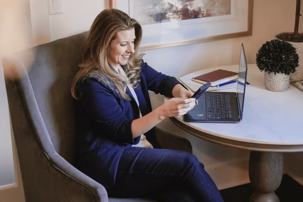 A woman in a blue blazer sits at a marble table using a phone, with a laptop and a notepad nearby. A potted plant decorates the space.