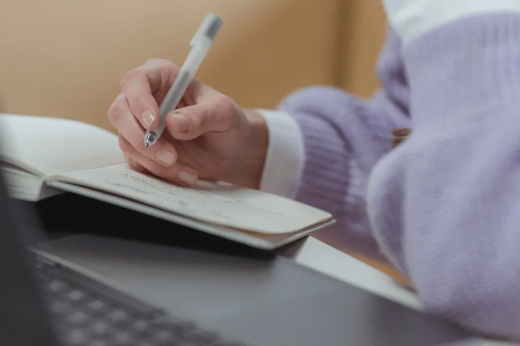 Person in a purple sweater jotting down notes in a planner with a pen.