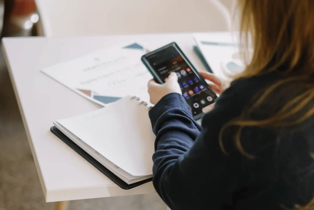 A person with long hair holds a smartphone while sitting at a table with a notepad and documents.