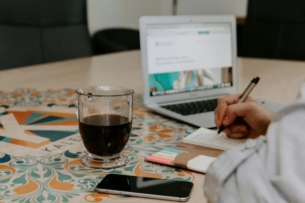 Person taking notes beside a laptop and coffee.