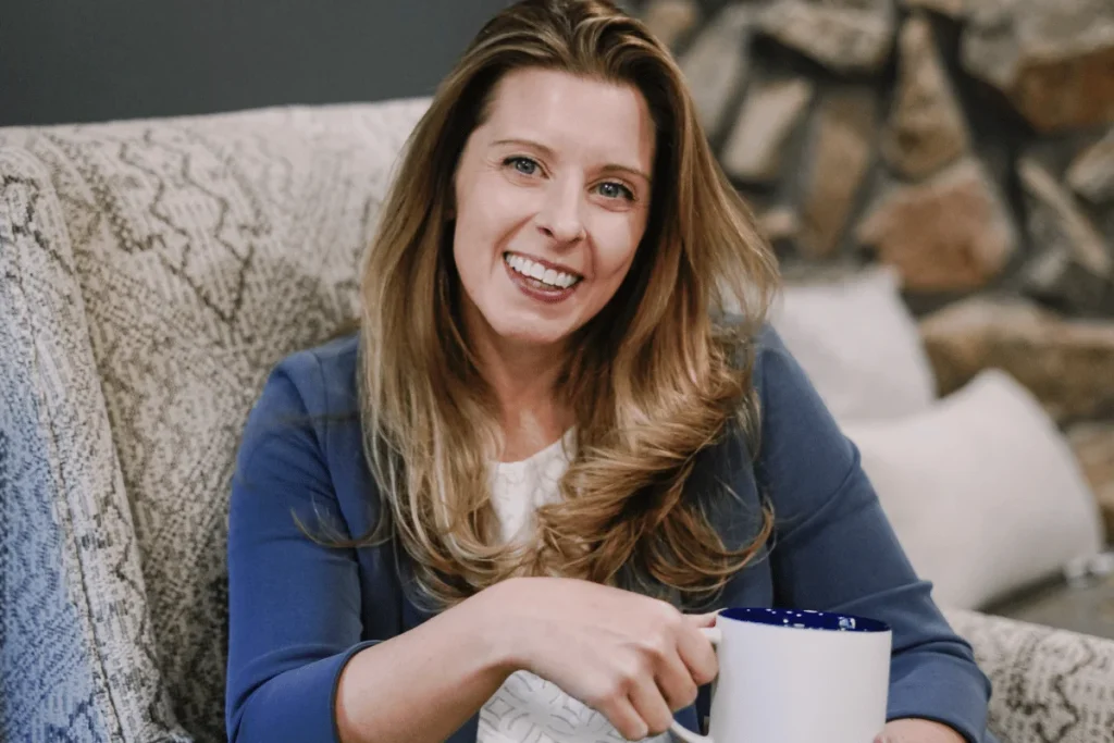 Nora Sudduth with long, wavy hair holds a white mug while seated on a patterned chair, with a stone wall in the background.