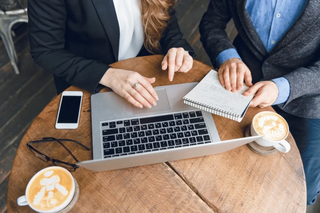 Two people collaborating at a café, using a laptop and notepad, with coffee cups on a wooden table. A phone and glasses are nearby.