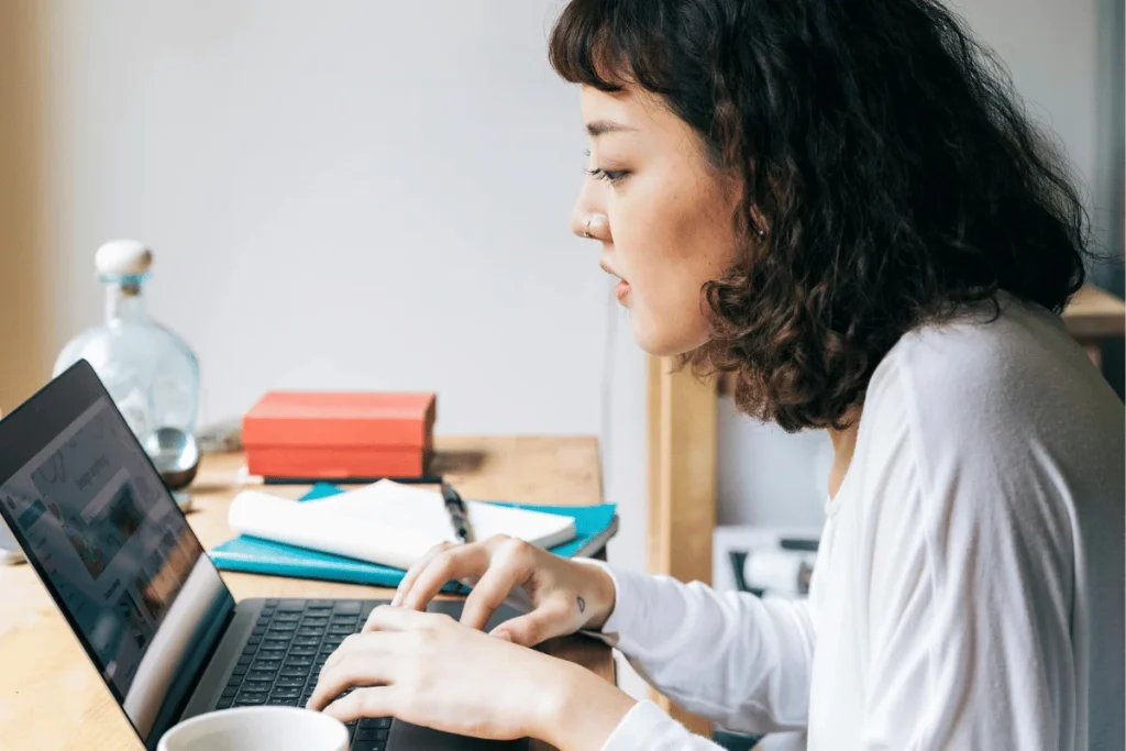 Focused lady using a laptop for research with books and coffee on her desk.