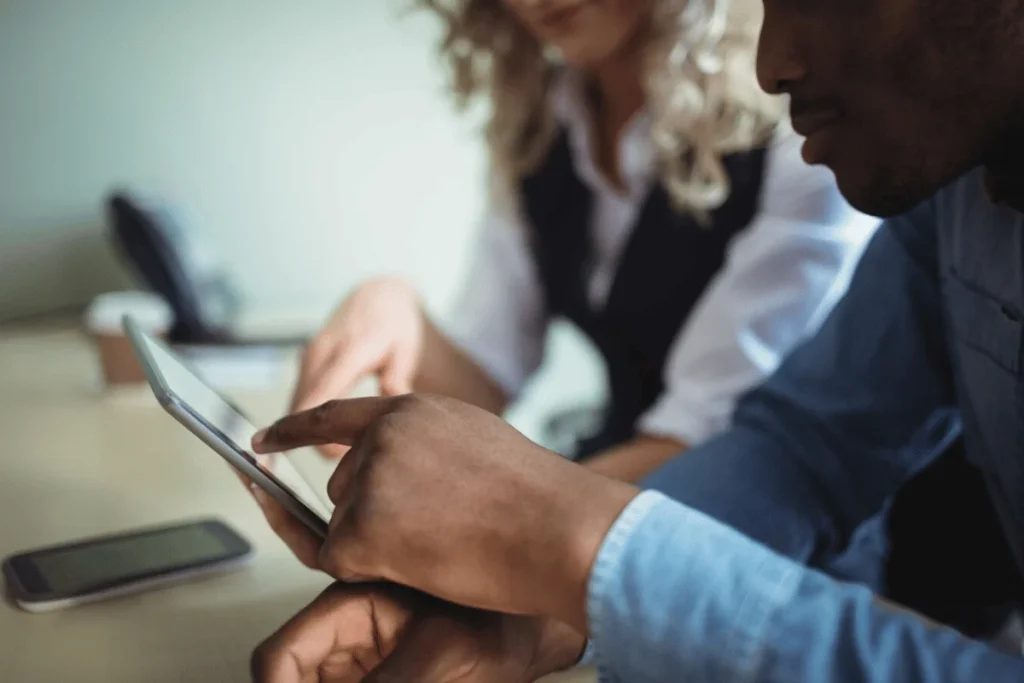 Two individuals collaborate over a smartphone on a table, with one pointing at the screen while the other observes.