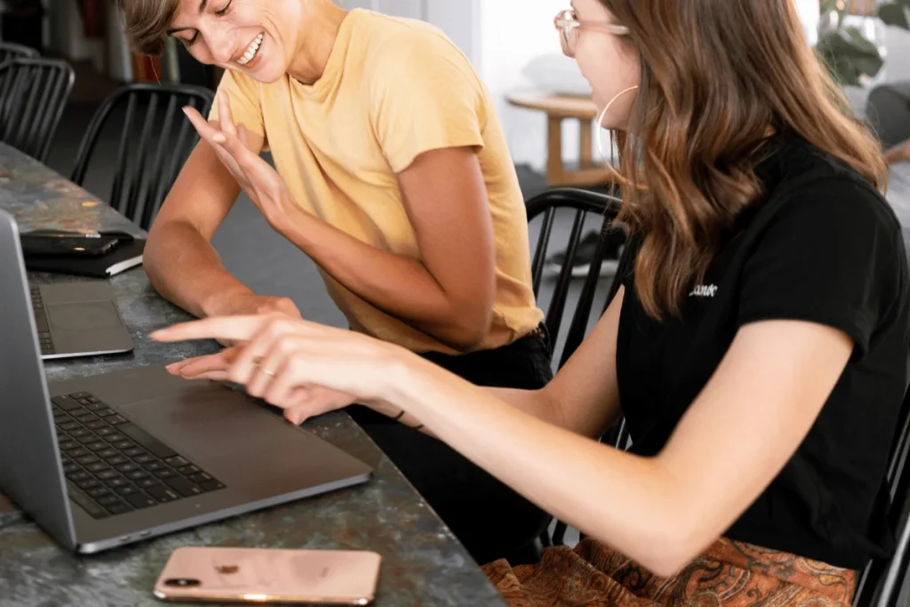 Two people sit at a table using laptops, smiling and interacting with each other. A smartphone and other devices are also on the table.