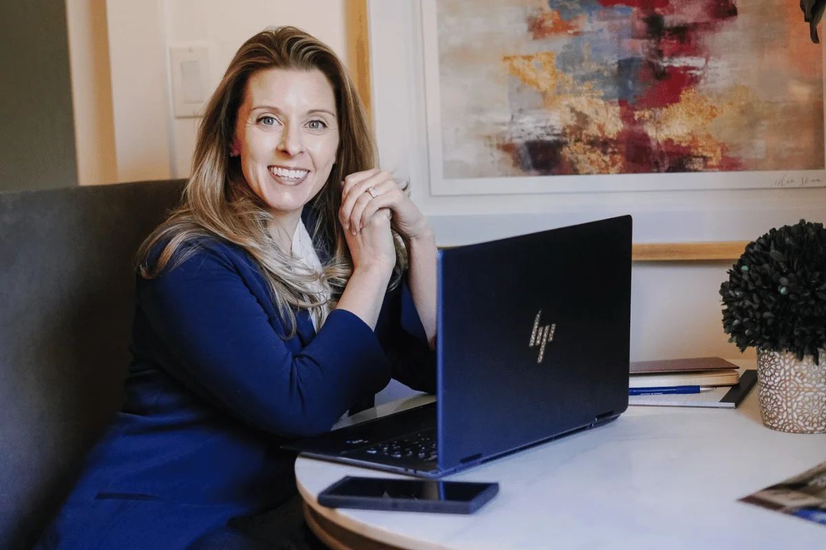 A business-focused Nora Sudduth smiles while working on her laptop in a stylish room.