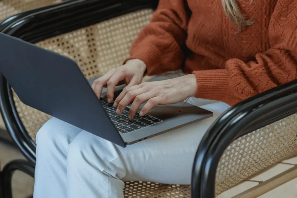 Individual in orange sweater using laptop on rattan chair.