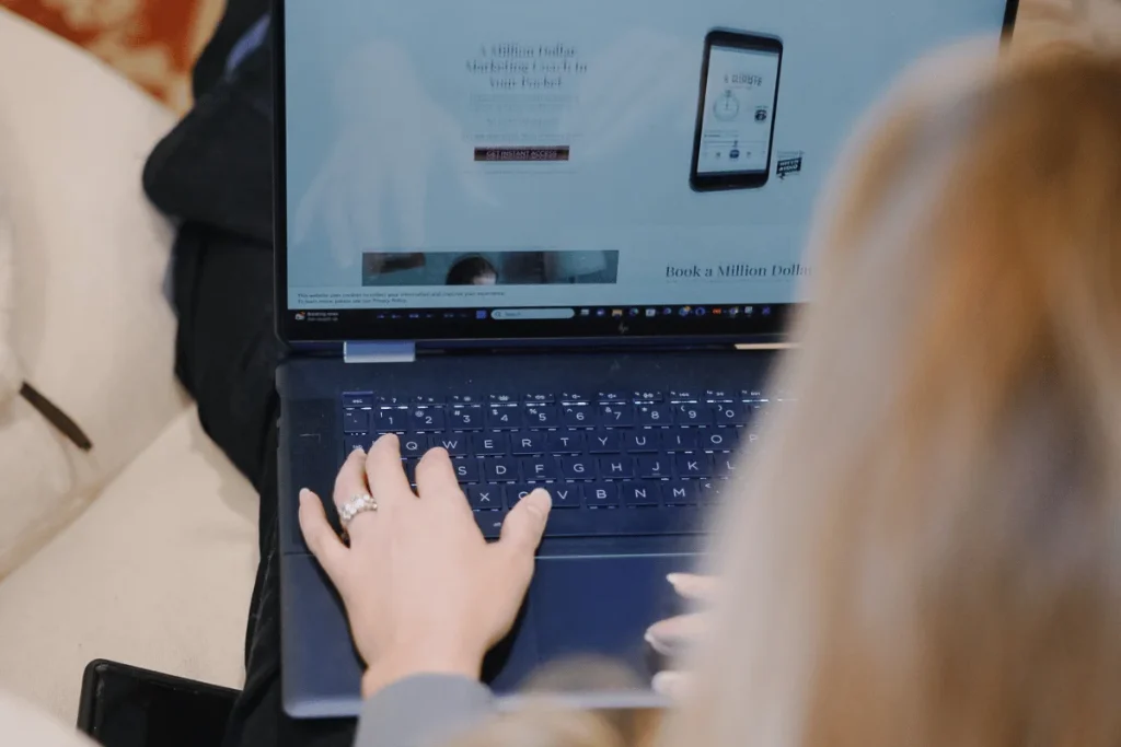 A person with long hair types on a laptop displaying a marketing webpage. The screen shows a smartphone image and text. The setting is relaxed.