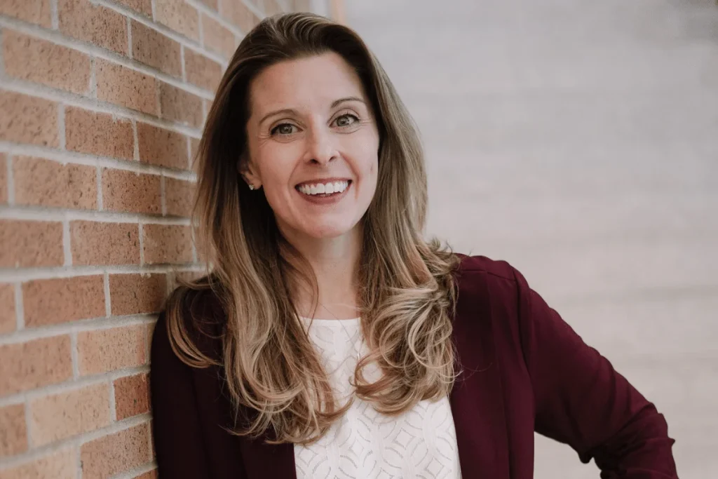 Warmly smiling Nora Sudduth leaning against a brick wall.