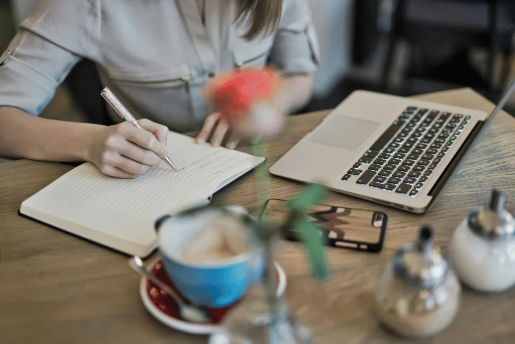 Freelancer making notes in a journal with a laptop, smartphone, and coffee on the table.