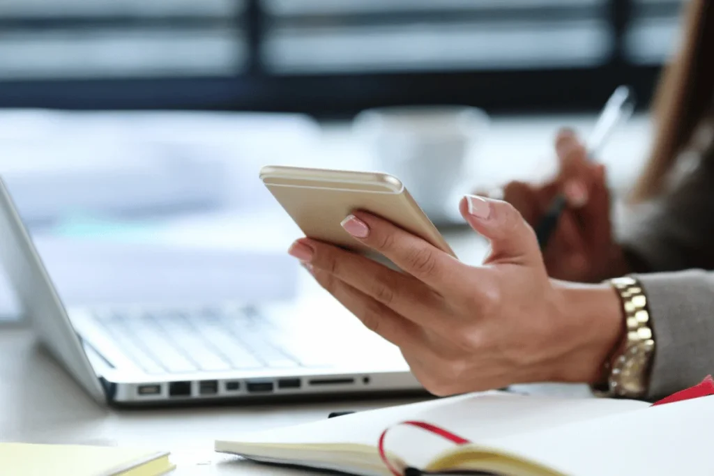 A close-up of a person's hand holding a smartphone, with a laptop and notepad in the background, suggesting a work environment.