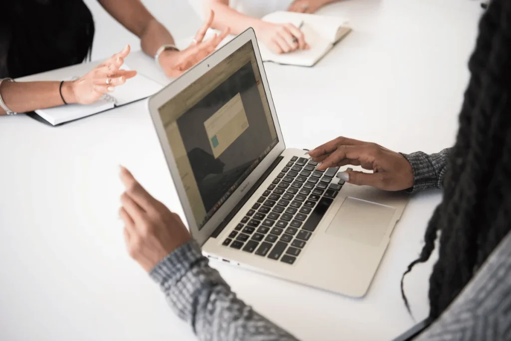 A person types on a laptop at a meeting table while others engage with gestures and notebooks, emphasizing teamwork and collaboration.