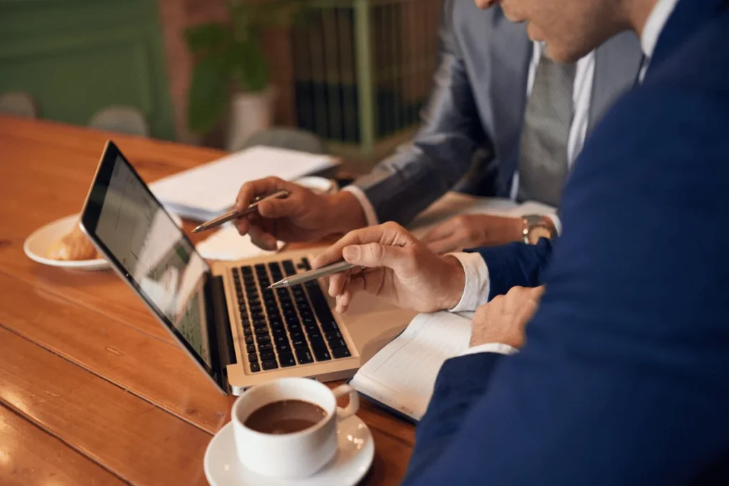 Two professionals collaborate over a laptop, pointing at the screen, with a notebook and a cup of coffee on the wooden table.