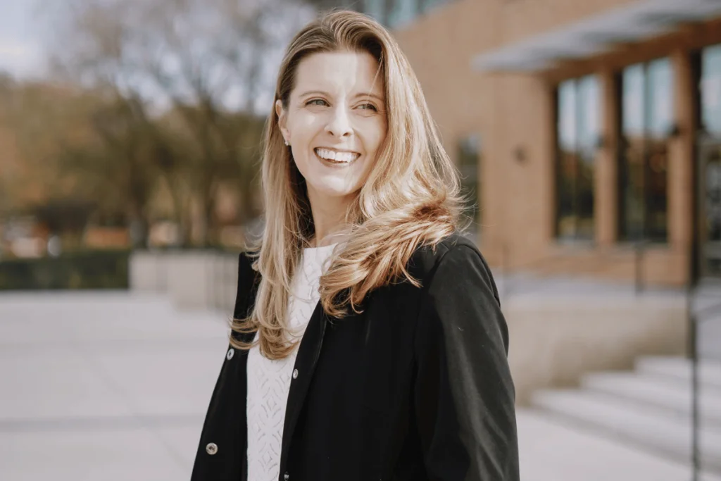Nora Sudduth in black coat, campus building backdrop.