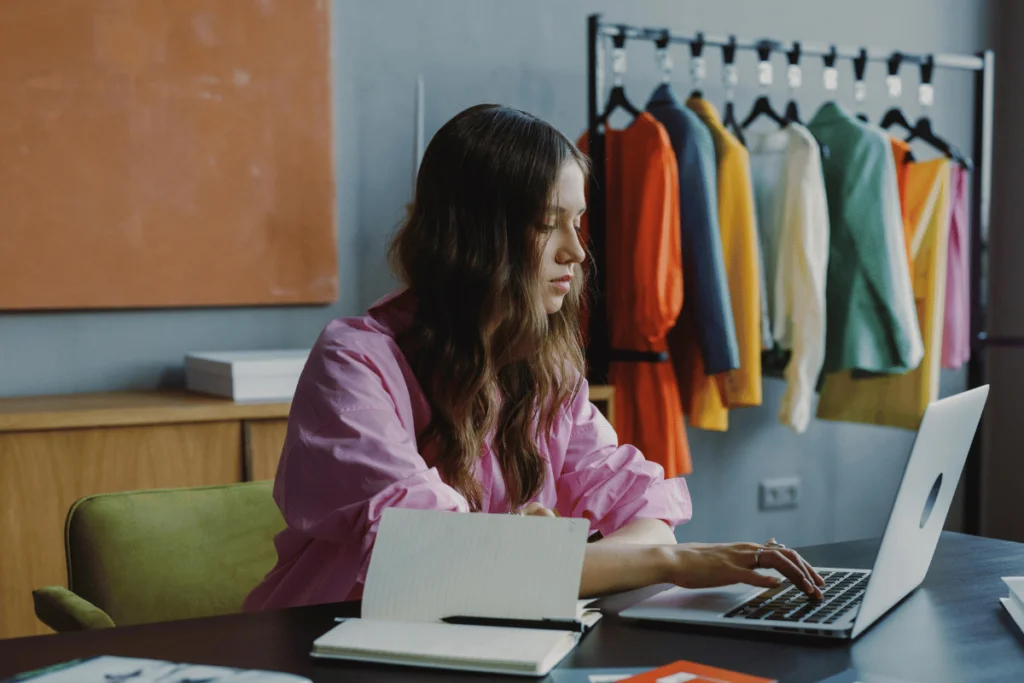 Woman in pink shirt focused on laptop in a fashion studio with clothes rack.
