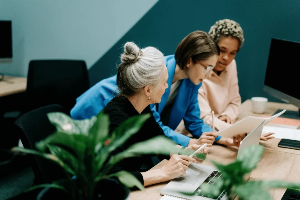 Three diverse colleagues collaborate at a desk with laptops and documents, conveying teamwork and focus. A green plant adds a touch of nature.