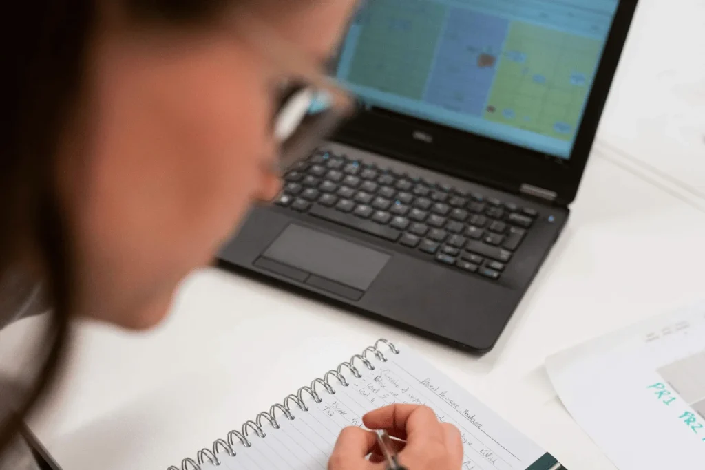 Executive assistant taking notes while analyzing data on laptop.