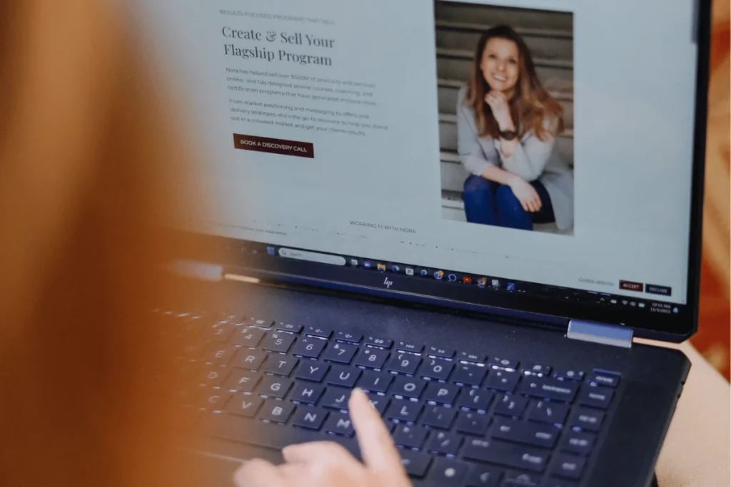 A person uses a laptop displaying a webpage titled "Create & Sell Your Flagship Program" with a photo of a smiling woman. The scene is focused and professional.