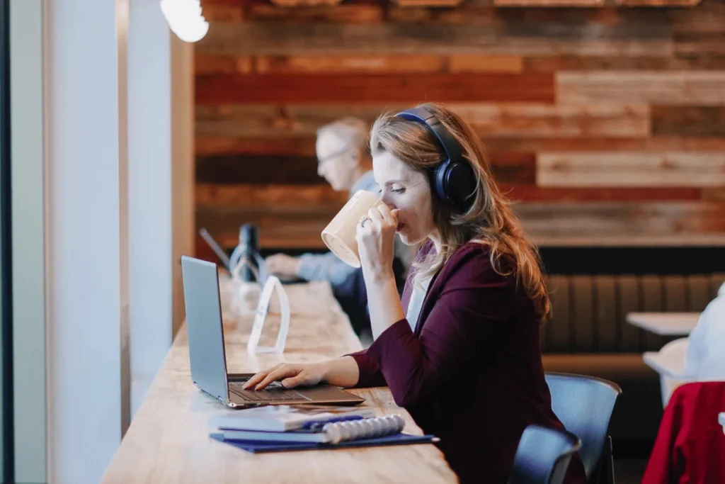 Nora Sudduth wearing headphones sits at a wooden table, typing on a laptop while holding a drink. The background features warm wooden paneling.