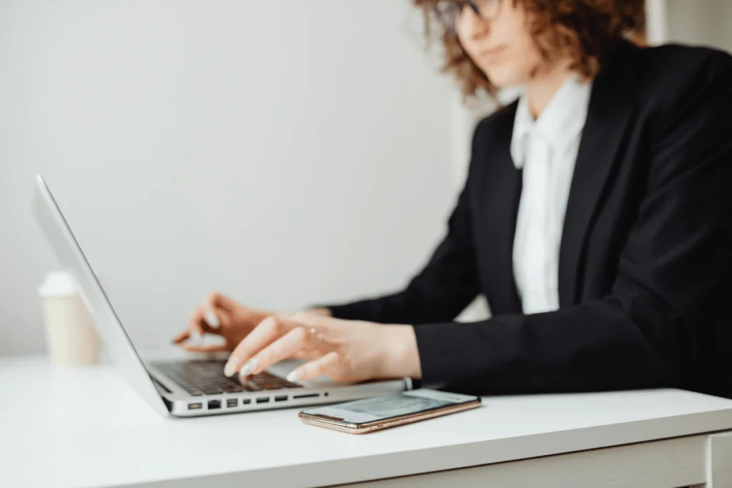 Professional in a blazer typing on a laptop with a smartphone beside her.