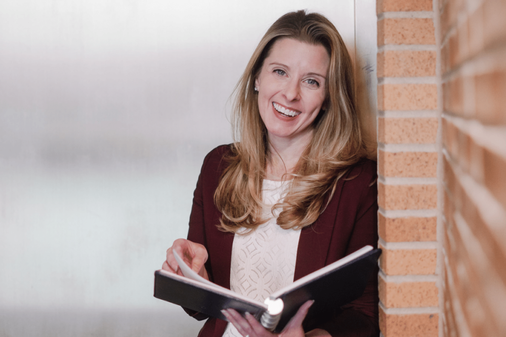 Nora Sudduth smiling confidently while holding an open notebook against a brick wall.