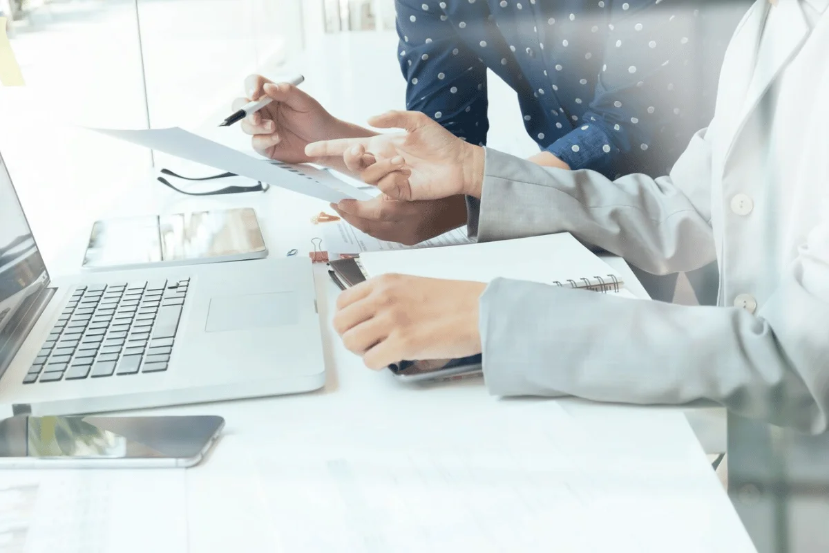 Two focused individuals discusses documents over a laptop, gesturing and writing notes in a collaborative office setting.