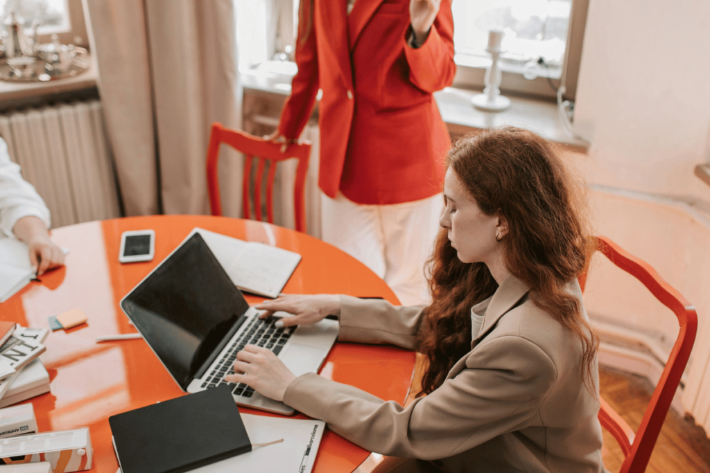 An elegant woman in a red suit conversing with a woman on a laptop.
