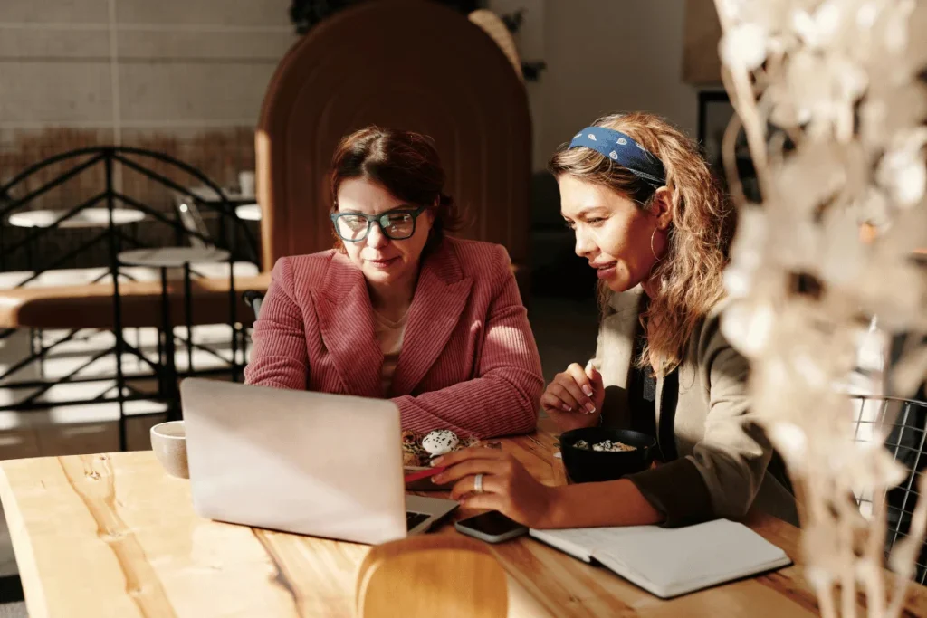 Two individuals sit at a wooden table in a cozy café, discussing over a laptop, with snacks and a notebook beside them.