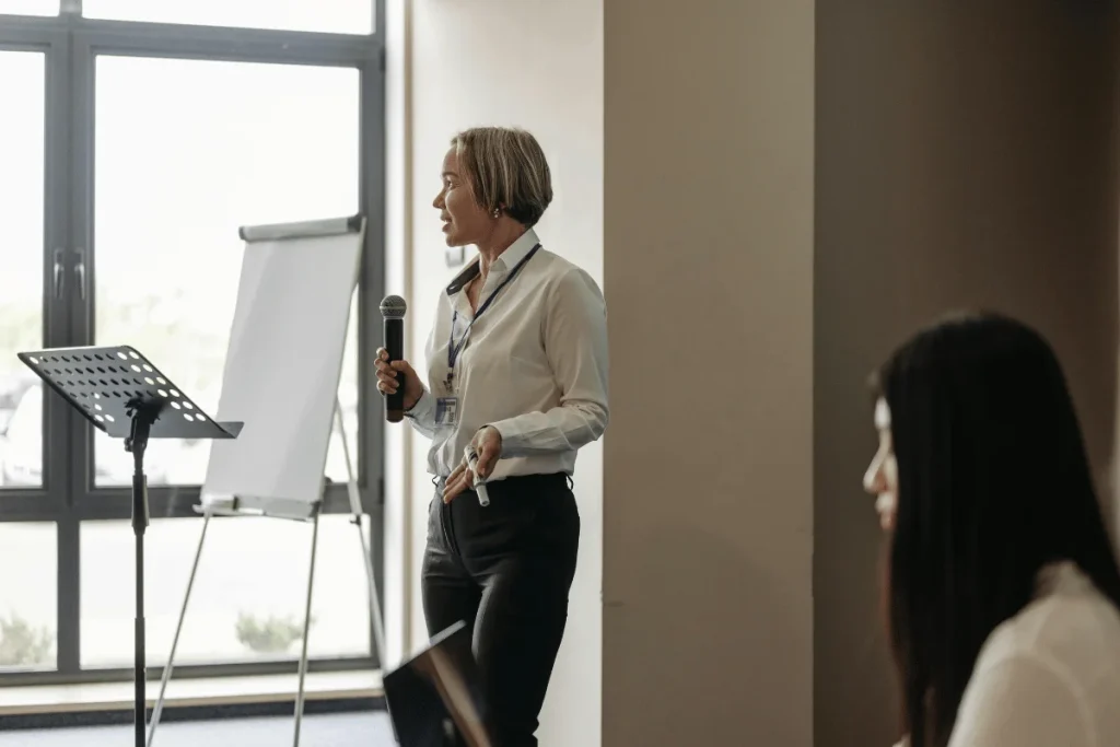A speaker stands in front of a flip chart, holding a microphone and gesturing while addressing an audience in a bright conference room.