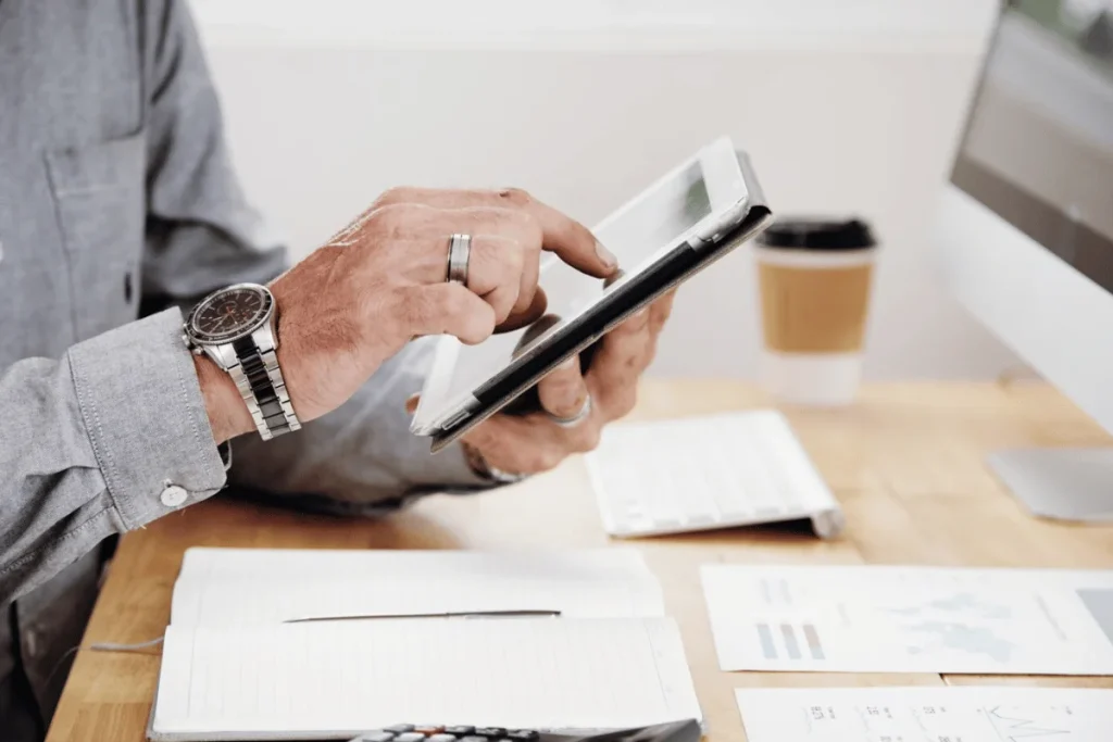 A person in a gray shirt uses a tablet at a wooden desk, surrounded by papers, a notebook, a calculator, and a coffee cup, conveying a productive work setting.