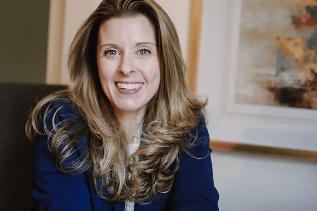 An inviting smile from Nora Sudduth, dressed in professional attire against an artful backdrop.