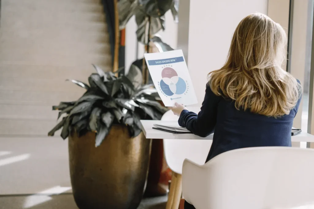 A woman sits at a table studying a document with sales data, beside a large potted plant, in a modern office space.