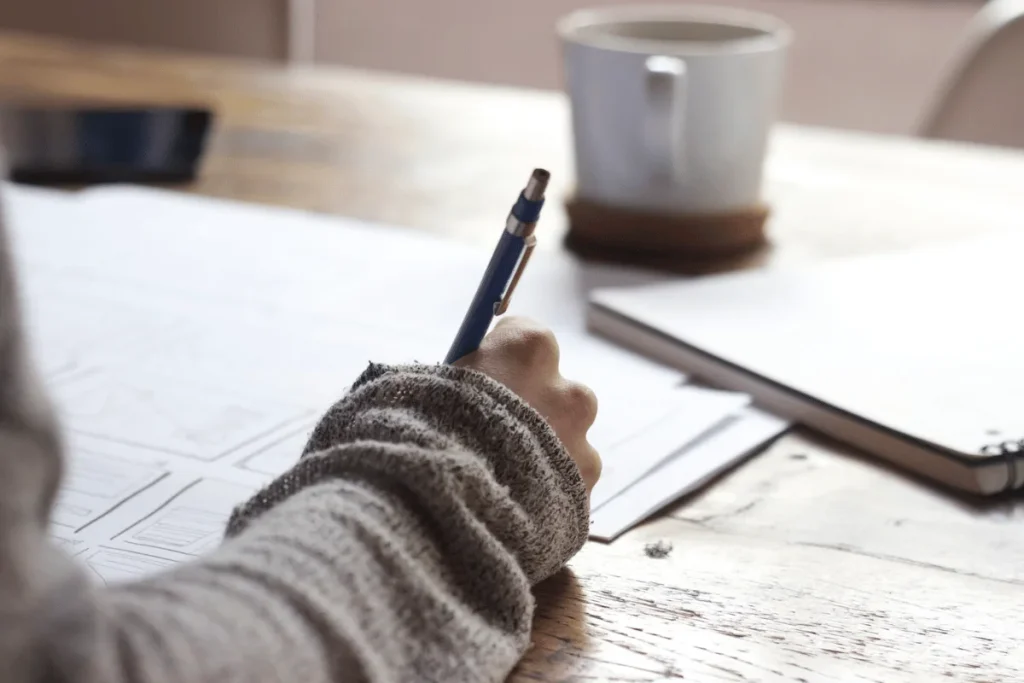 Close-up of a hand writing on architectural blueprints with a pen, coffee mug in the background.