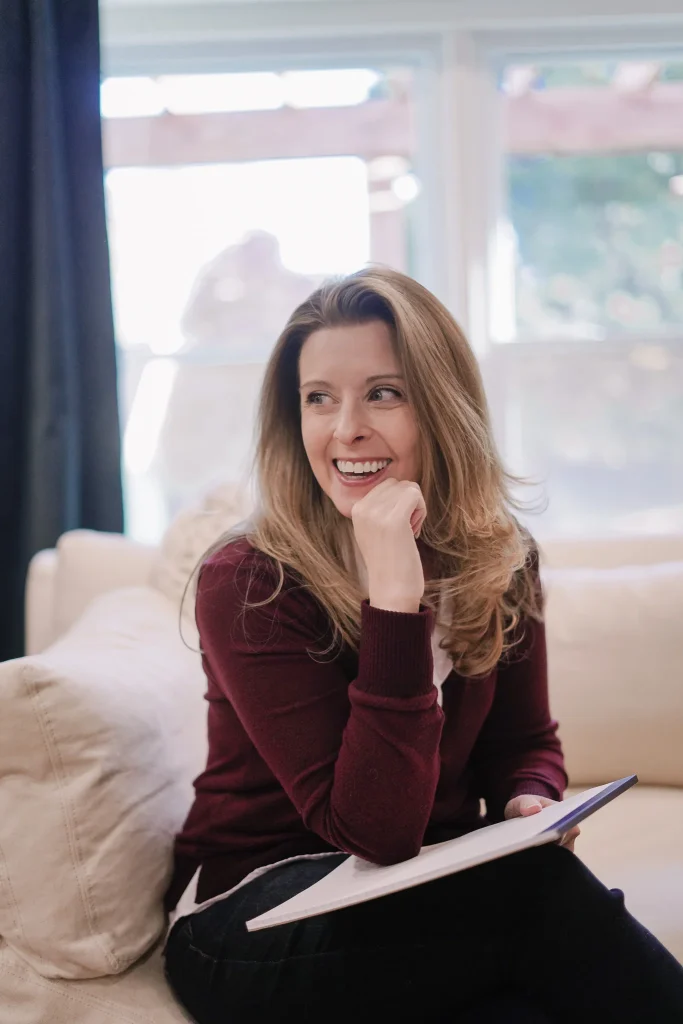 Nora wearing a burgundy sweater, smiling while holding a notebook. Bright window in the background. Relaxed and cheerful mood.