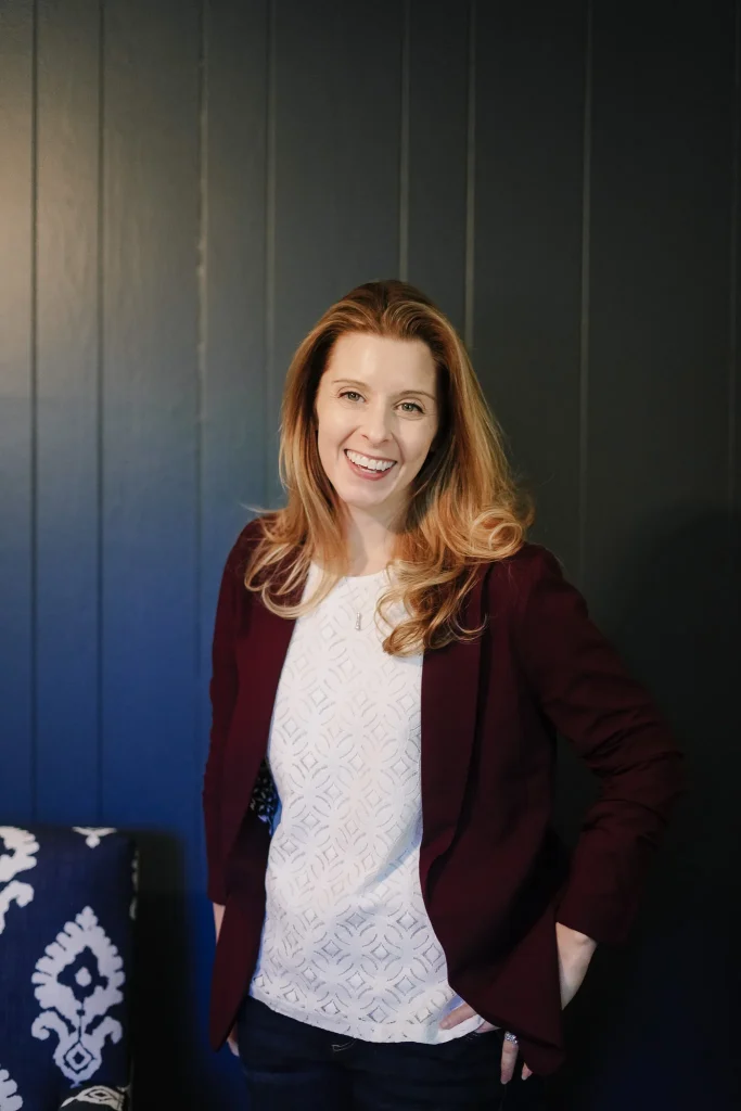 Nora with long hair wearing a maroon blazer and white patterned top, standing with hands in pockets against a dark paneled wall, exuding warmth and confidence.