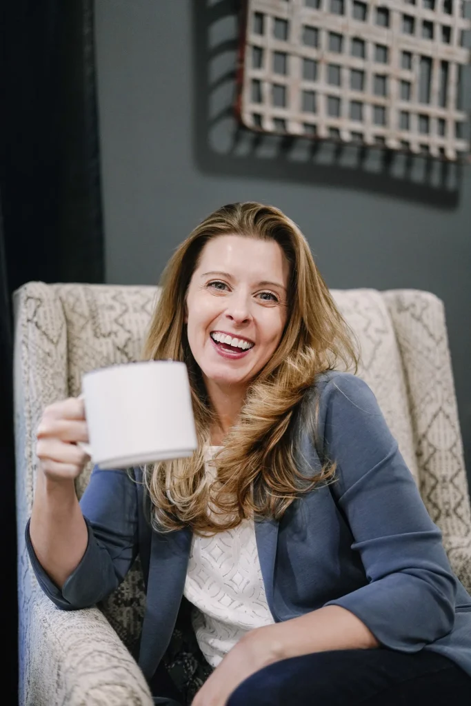 Nora Sudduth in a patterned armchair, holding a white mug.