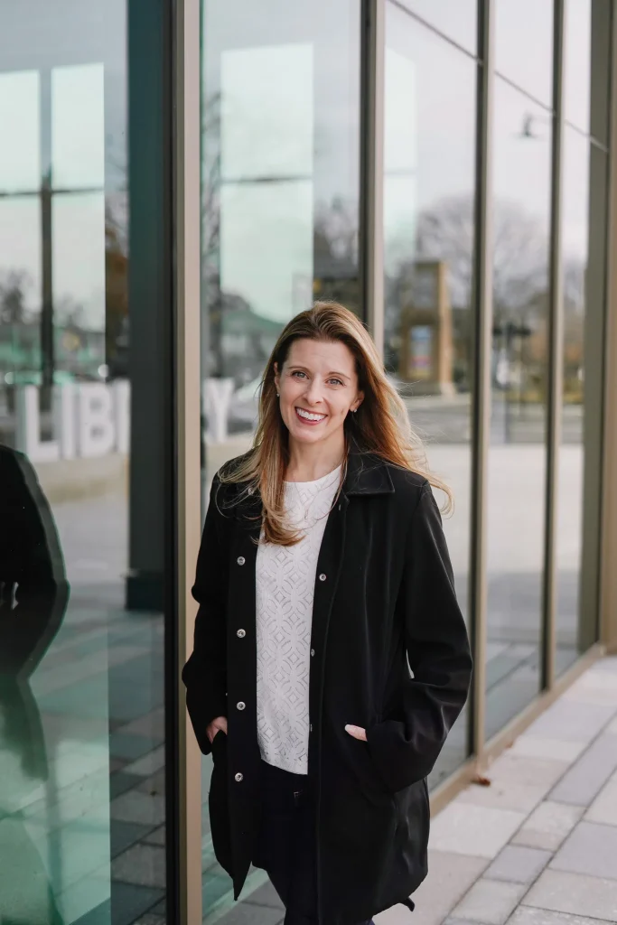Nora a black coat smiles while standing outside a modern building with glass windows, reflecting an urban setting and a calm, pleasant atmosphere.