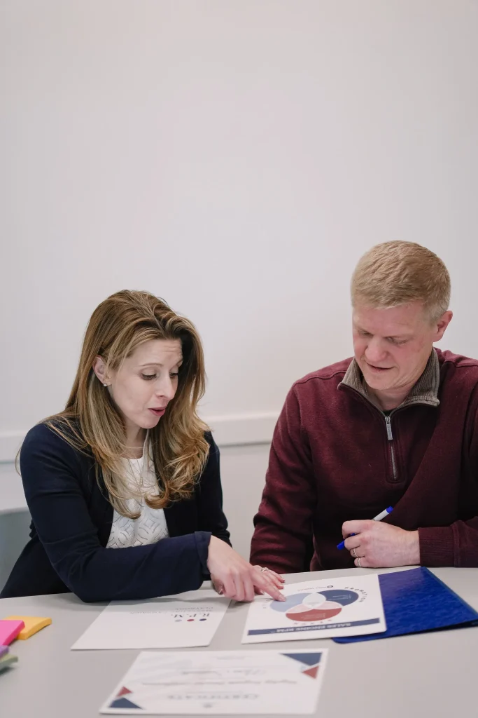 A woman and a man sit at a table, discussing a document with a colorful chart. The woman points at the chart. Both appear focused and engaged.