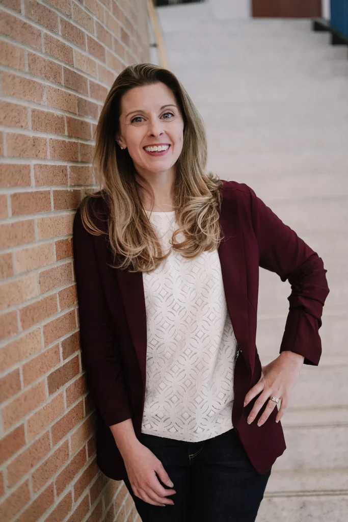 Nora in a maroon blazer smiling while leaning against a brick wall.
