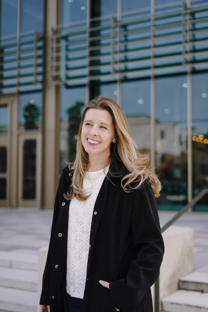 Nora wearing a black coat and white sweater stands outside a modern glass building, conveying confidence and warmth.