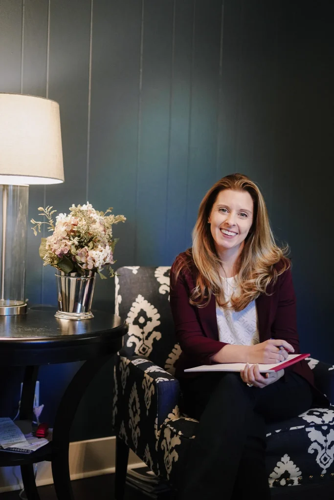 Nora sitting in patterned chair with notebook beside lamp and flowers.