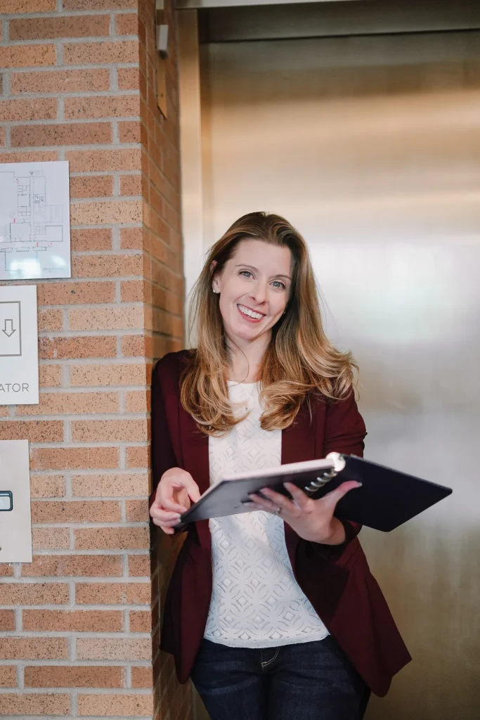 Nora holding a notebook while standing near an elevator.
