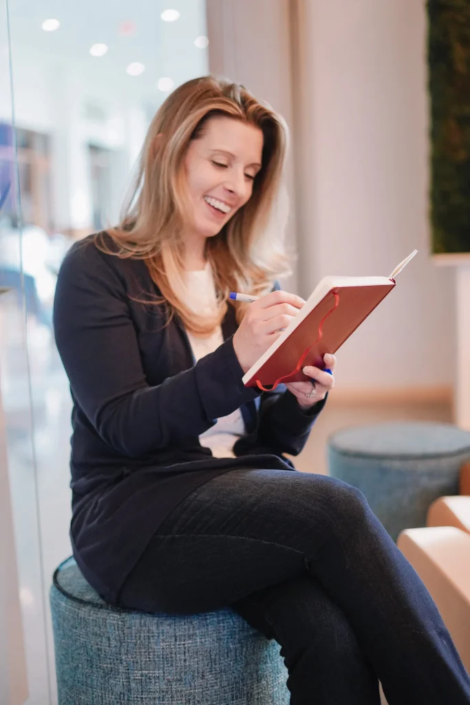Nora sitting on a stool, smiling while writing in a red notebook. She appears relaxed and happy in a modern, softly lit environment.