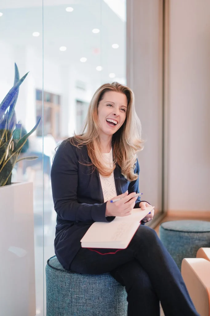Nora Sudduth with long blonde hair smiles and writes in a notebook while sitting on a stool. She is in a modern, bright setting with a plant beside her.