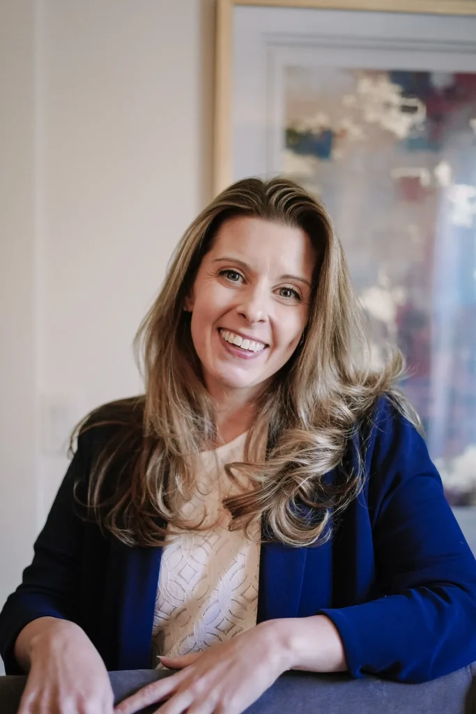 Smiling woman with long hair in a blue blazer and white top, seated indoors with abstract art in the background, conveying a friendly and approachable tone.