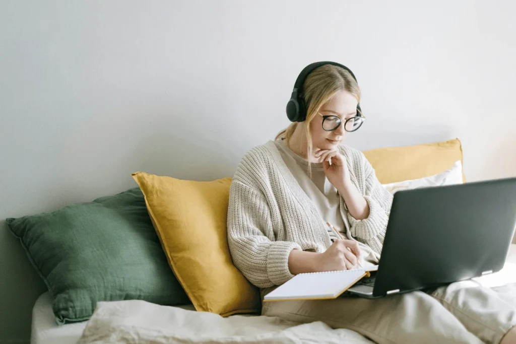A woman studies on her bed, wearing headphones and using a laptop alongside a notebook.