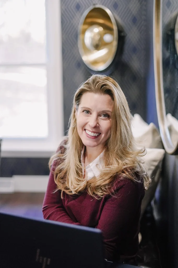 Smiling Nora in a gray blazer and jeans sits on indoor stairs in a relaxed professional portrait.