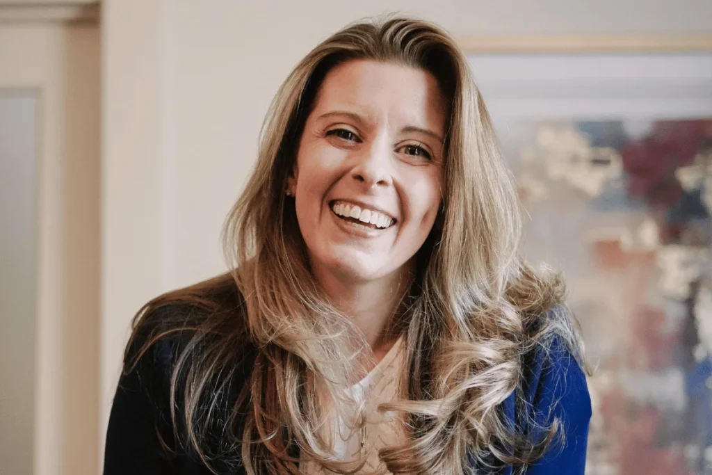 Nora Sudduth in blue jacket portrait, featuring casual professional style against an abstract indoor background.