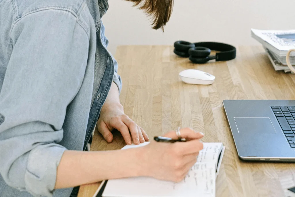 Workspace setup featuring a person writing in a journal, with a laptop, wireless mouse, and headphones.