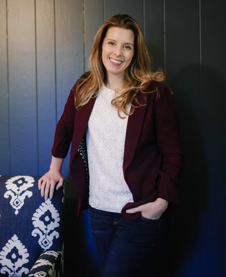 Nora Sudduth stands against a dark paneled wall, wearing a burgundy blazer and white patterned shirt.